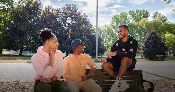 Photograph of three people sitting and chatting outdoors on a park bench with trees and a pathway in the background. Two individuals sit on the bench while one sits on the armrest, all smiling and engaged in conversation on a sunny day.