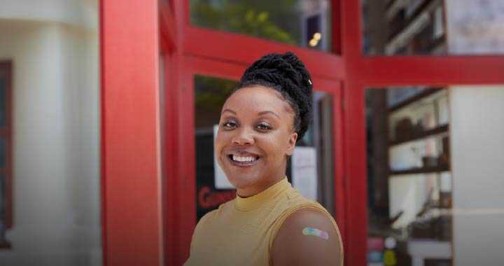 Photograph of a smiling woman wearing a yellow sleeveless top standing outside a building with red-framed glass doors. A colorful adhesive bandage is visible on her upper arm, indicating she recently received a vaccination.