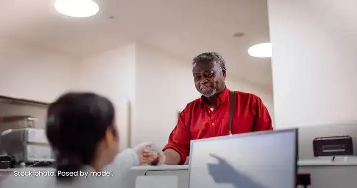 Photograph depicting a customer-service interaction between a smiling Black male employee in a red shirt and a customer at a counter.