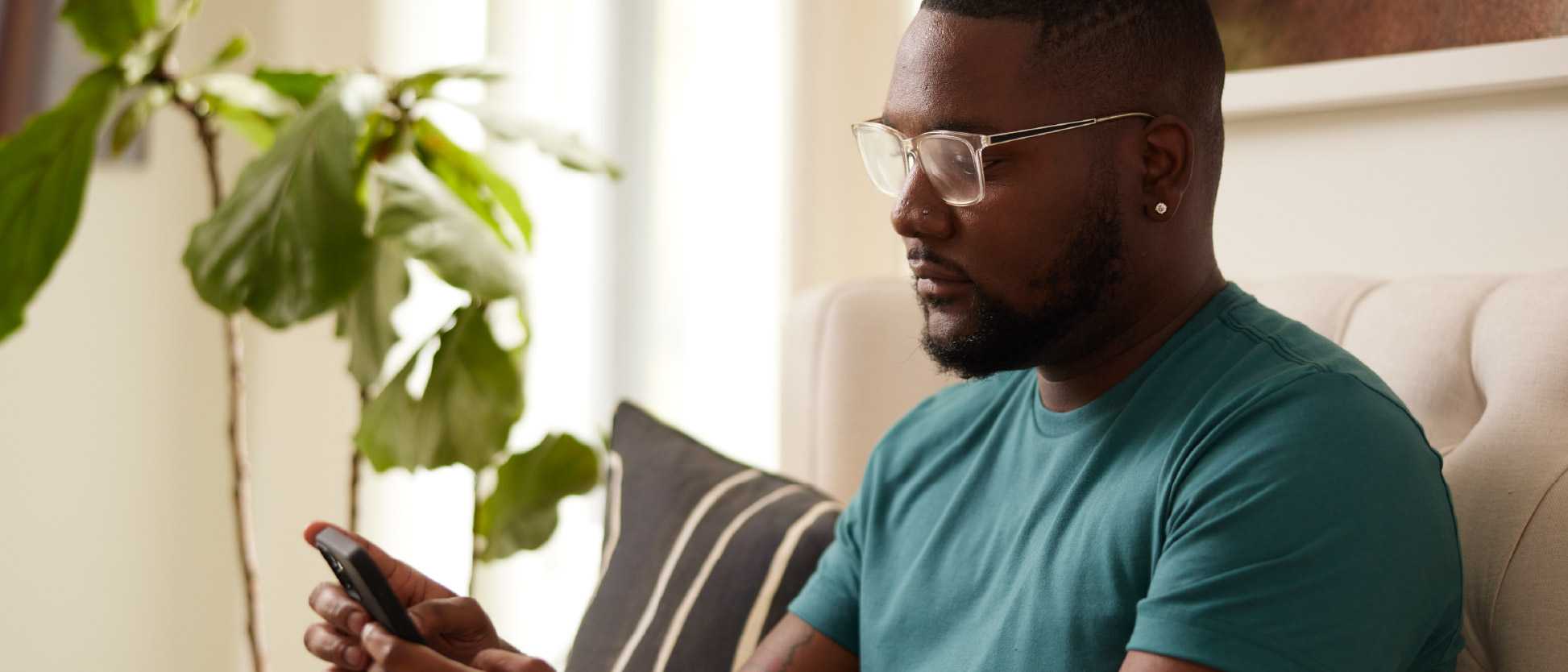 Photograph of a man wearing glasses and a teal shirt sitting on a couch while looking at his smartphone. The setting includes a large green plant and a pillow with black and white stripes, suggesting a relaxed indoor environment.