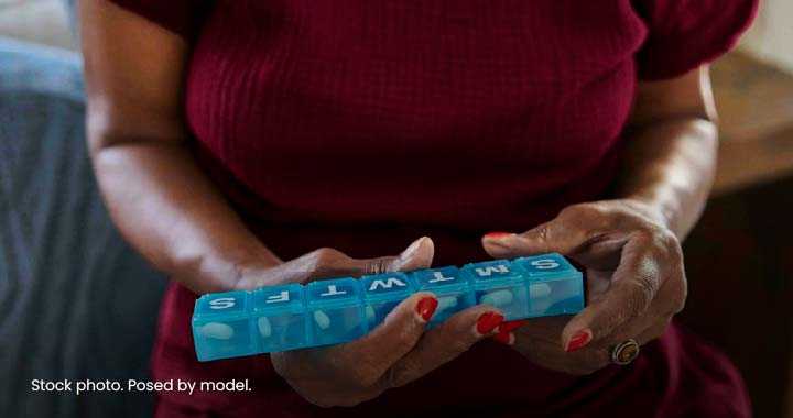 Photograph showing a person holding a blue weekly pill organizer with compartments labeled S, M, T, W, T, F, S containing white pills. The image highlights medication management and organization, with focus on hands and pillbox against a blurred background.