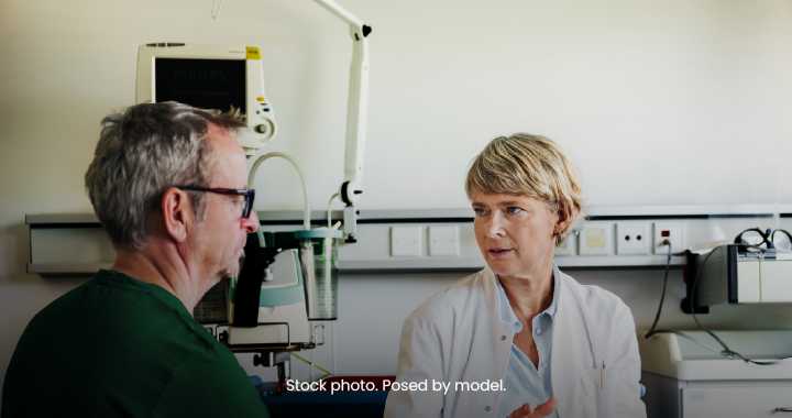 Photograph of a medical consultation showing a female doctor in a white coat speaking with a male patient wearing glasses and a green shirt. Setting includes medical equipment and a clinical environment, highlighting patient-doctor communication and healthcare context.