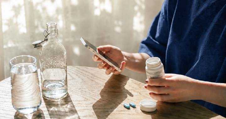 Photograph showing a person preparing to take medication beside a glass and bottle of water. Open pill container, white tablets, blue capsules, smartphone, and sunlit wooden table highlight medication use and hydration.