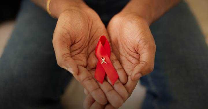 Photograph showing a pair of hands gently holding a red ribbon with a small white awareness ribbon pinned on it. The red ribbon symbolizes HIV/AIDS awareness and support, emphasizing care and solidarity.